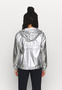 Silver lättviktig jacka med en glänsande, rynkad yta, med en fast huva och elastiska ärmslut. Ingen synlig logotyp.