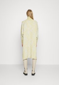 Gul stickad klänning med vågmönster, hög krage, långa ärmar och avslappnad passform, matchad med krämfärgade höga stövlar.