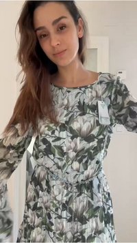 Γυναίκα με μακριά καστανά μαλλιά, που φοράει πράσινο και λευκό φόρεμα με λουλούδια, ζώνη αλυσίδα και ορατή ετικέτα της μάρκας Guess.