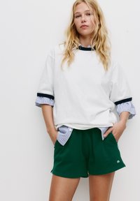 Übergrößes weißes Baumwoll-T-Shirt mit navyfarbenen Bündchen über einem blau-weiß gestreiften Hemd, kombiniert mit grünen Shorts mit Seitentaschen.