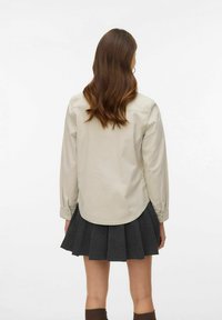 Beżowa koszula z długim rękawem o zaokrąglonym dole, zestawiona z plisowaną, ciemnoszarą spódnicą. Miękka tkanina o gładkiej fakturze i mankiety na guziki.