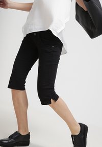 Černé zkrácené kalhoty s přiléhavým střihem, předními kapsami a jemnými bočními rozparky na lemech. Nošené s černými botami a bílým topem.