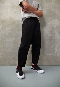 Personne debout, les bras croisés, portant un t-shirt gris, un pantalon noir et des baskets noires, blanches et rouges, devant un mur en béton gris.