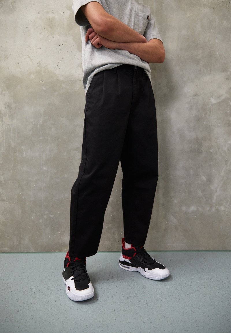 Personne debout, les bras croisés, portant un t-shirt gris, un pantalon noir et des baskets noires, blanches et rouges, devant un mur en béton gris.