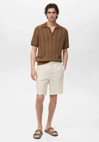 Brunstribet kortærmet polo med V-udskæring, kombineret med cremefarvede shorts med snoretræk og brune sandaler, mod en neutral baggrund.