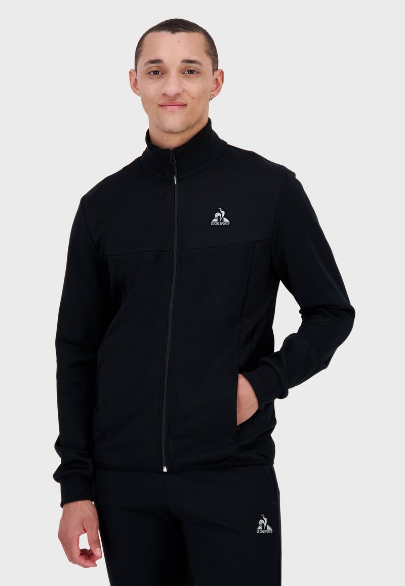 Giacca nera con zip realizzata in tessuto morbido, dotata di un colletto alto, un pannello in rete e un logo sul petto. Design funzionale con una vestibilità comoda.