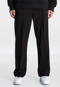 Schwarze, locker sitzende Sweatpants mit glatter Oberfläche, mit vorderen Falten und einem dezenten Logodetail an der Taille. Kombiniert mit weißen Sneakers.