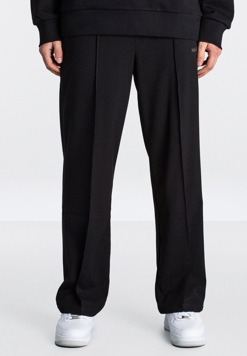 Schwarze, locker sitzende Sweatpants mit glatter Oberfläche, mit vorderen Falten und einem dezenten Logodetail an der Taille. Kombiniert mit weißen Sneakers.