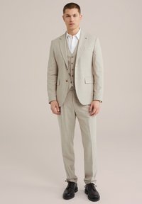 Beige Dreiteiler mit kariertem Muster, tailliertem Schnitt, einreihigem Sacko und braunen Knöpfen. Kombiniert mit einem weißen Hemd und schwarzen Schuhen.