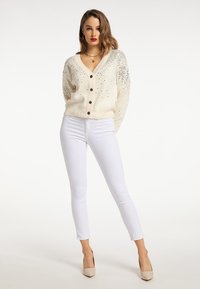 Donna che indossa un cardigan crema con bottoni e paillettes, pantaloni bianchi attillati, décolleté beige, con le mani dietro la schiena su uno sfondo bianco.