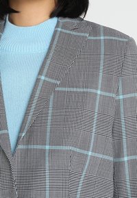 Szary blazer w kratę z jasnoniebieskimi akcentami, z teksturowanym wzorem i usztywnionymi klapami. Noszony na jasnoniebieskim swetrze.