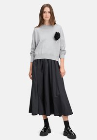Grau gestrickter Pullover mit einem gerippten Rundhalsausschnitt, verziert mit einem schwarzen Stoffblüten-Akzent. Kombiniert mit einem schwarzen A-Linien-Rock und schwarzen Plateau-Schuhen.