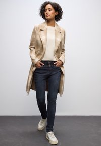 Abrigo largo a cuadros beige sobre una blusa crema, combinado con jeans oscuros ajustados y zapatillas crema. Detalles destacados incluyen un cuello estructurado y bolsillos.