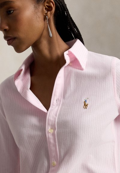Giovane donna con capelli intrecciati indossa un orecchino a goccia d'argento e una camicia rosa chiaro con bottoni con un piccolo logo di cavallo e cavaliere sul petto.
