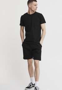 Sort kortærmet t-shirt med en brystlomme, parret med sorte shorts. Modellen bærer sorte og hvide sneakers med hvide ankelsokker.