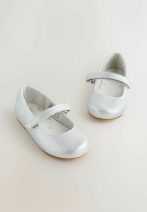 Pár strieborných trblietavých detských topánok v štýle Mary Jane s suchými zipsami a svetlohnedými podrážkami na neutrálom pozadí.