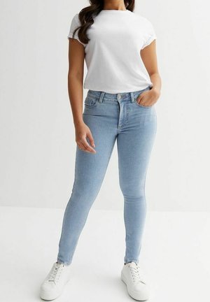 Femme portant un jean skinny bleu clair, un t-shirt blanc à manches courtes et des baskets blanches, debout avec une main dans la poche.