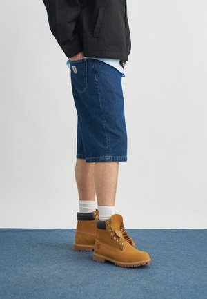 Személy kék farmer rövidnadrágot, fehér zoknit, barna munkacipőt és fekete dzsekit viselve, kék szőnyeggel borított padlón áll.