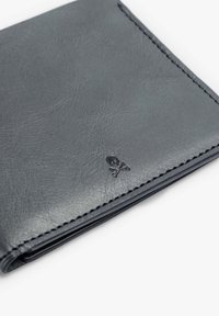 Cartera de cuero gris con costuras visibles y un pequeño logo de calavera y huesos cruzados. La superficie tiene una textura suave y un brillo sutil.