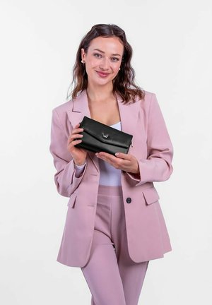 Černá kožená peněženka s překlopným víkem a kovovým akcentem, držena v rukou. Modelka v růžovém obleku stojí na bílém pozadí.