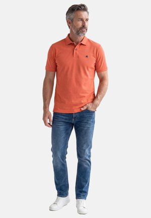 Mand med gråt hår og skæg iført en orange polo, blå jeans og hvide sneakers, stående med den ene hånd i lommen og kiggede til siden.