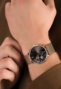 Montre ronde avec un cadran noir, des accents argentés et un bracelet en maille dorée. Comprend trois sous-cadrans de chronographe et des chiffres surdimensionnés.
