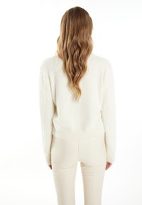 Weißer, flauschiger Pullover mit rundem Halsausschnitt und geripptem Saum. Der Stoff wirkt weich und strukturiert, kombiniert mit hellen, figurschmeichelnden Hosen.