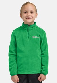 Bambino sorridente con capelli biondi che indossa un maglione in pile verde brillante di Jack Wolfskin e pantaloni neri, in piedi su uno sfondo semplice.