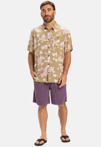 Kurzarm, beiges Hemd mit Knöpfen und rosa-weißen Blumenmustern, kombiniert mit lila Shorts und schwarzen Sandalen.