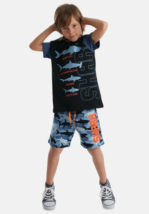 Schwarzes und blaues T-Shirt mit Haifischmotiv und Text, kombiniert mit blauen Camo-Shorts, die Haifischgrafiken und einen orangefarbenen Akzent zeigen. Graue Sneakers.