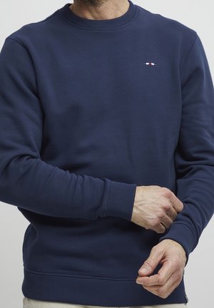 Mann richtet Ärmel eines marineblauen Sweatshirts mit Rundhalsausschnitt und kleinem rot-weiß-blauem Logo auf der Brust.