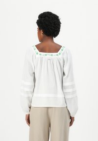 Witte blouse met groene bloemenborduring langs de vierkante halslijn. Lange pofmouwen met horizontale strepen. Textuur van katoen.