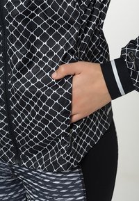 Hand in pocket of black jacket with white diamond pattern, black zipper, and reflector stripe on cuff; paired with black leggings with white dashes.