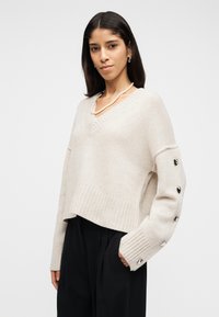 Femme portant un pull crème à col en V avec des boutons argentés sur la manche, un collier de perles, un pantalon noir, les mains dans les poches, sur fond blanc.