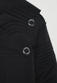 Black quilted jacket featuring a high collar, silver hardware accents, and distinct linear stitching patterns. Textured fabric with a smooth finish.