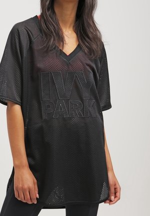 Vrouw draagt een losse zwarte mesh V-hals sportshirt met 'IVY PARK' geborduurd op de voorkant, staand tegen een effen achtergrond.