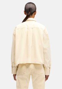 Giacca accorciata di colore crema, realizzata in tessuto resistente. Presenta una vestibilità comoda, una piega centrale sul retro e polsini con bottoni. Design semplice e pulito.