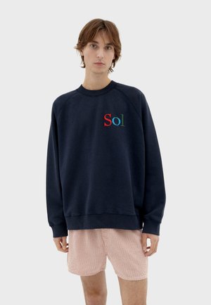 Granatowy sweatshirt z żebrowanym okrągłym dekoltem, z kolorowym haftem "Sol" na piersi i rękawami raglanowymi. W zestawie z szortami w paski.
