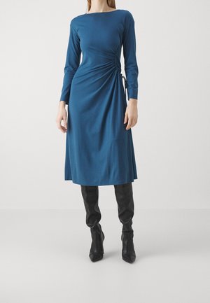 Teal long-sleeve dress with a side ruching detail, made of soft fabric. Paired with fitted black boots. Simple, elegant silhouette.