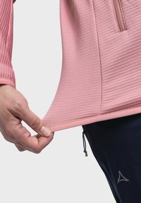 Top sportivo rosa chiaro con una texture a coste, dotato di colletto con zip e orlo regolabile, tenuto da una mano. Abbinato a pantaloni sportivi neri.