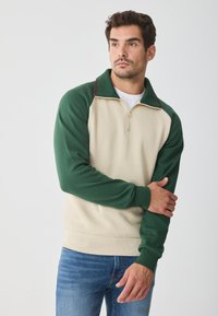Sudadera beige y verde con cremallera de cuarto, con mangas raglán, colores en contraste y tela texturizada. Llevada sobre una camiseta blanca.