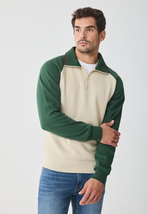 Sudadera beige y verde con cremallera de cuarto, con mangas raglán, colores en contraste y tela texturizada. Llevada sobre una camiseta blanca.