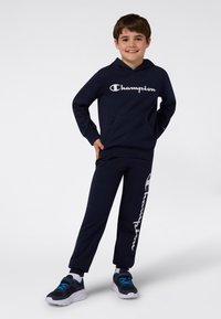 Navy bawełniana bluza z kapturem z białym logo "Champion", w komplecie z dopasowanymi dresami z bocznym logo. Ciemne buty sportowe z niebieskimi akcentami.