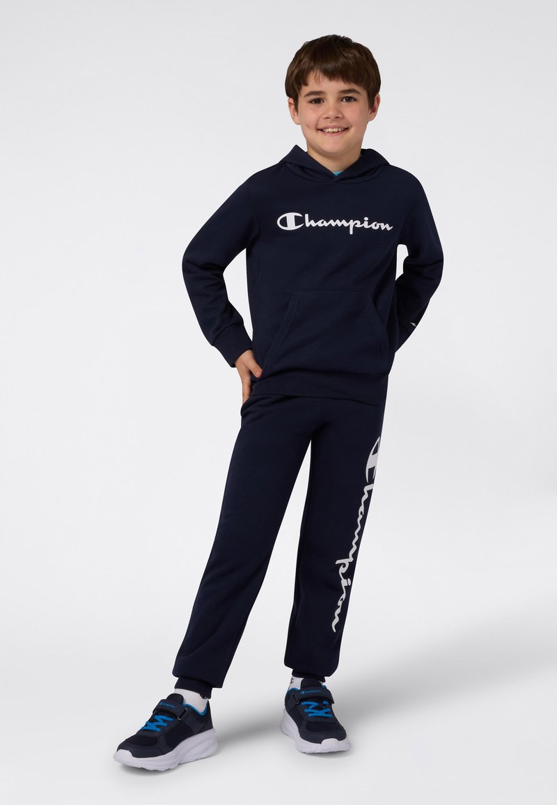 Navy bawełniana bluza z kapturem z białym logo "Champion", w komplecie z dopasowanymi dresami z bocznym logo. Ciemne buty sportowe z niebieskimi akcentami.