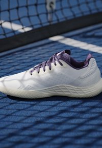 Valge ja lilla tennisesärk, mis puhkab sinisel väljakul musta võrgu lähedal, mis loob mustrilisi varje.