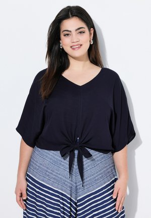 Námořnický modrý crop top s výstřihem do V a krátkými rukávy, s předním uzlem. V kombinaci s modrou sukní s vertikálním proužkem.