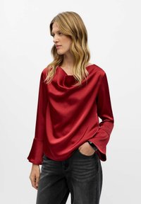 Röd satin topp med halsringning i kimonostil och trumpetärmar, kombinerad med mörka jeans, som visar en jämn textur och ett flödande mönster.
