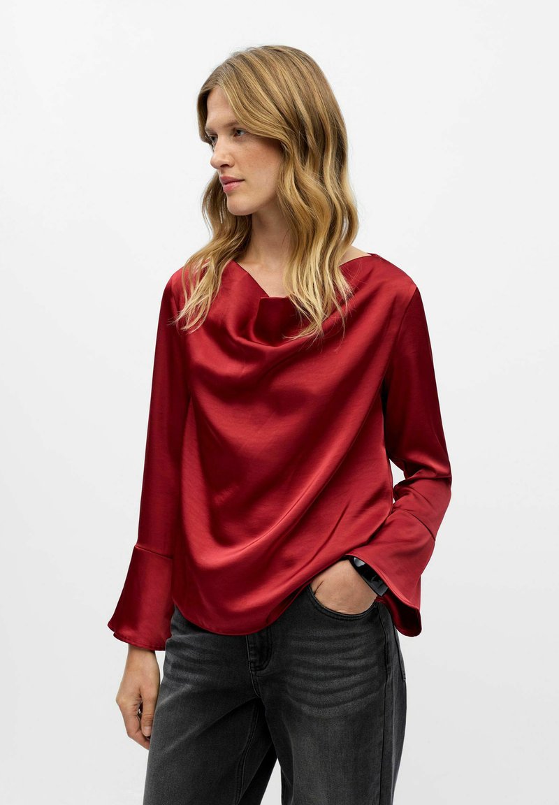 Röd satin topp med halsringning i kimonostil och trumpetärmar, kombinerad med mörka jeans, som visar en jämn textur och ett flödande mönster.