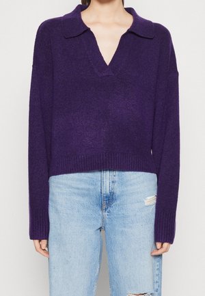 Lila stickad tröja med krage och V-ringning, med en avslappnad passform och ribbade ärmslut, ihop med ljusblå slitna jeans.