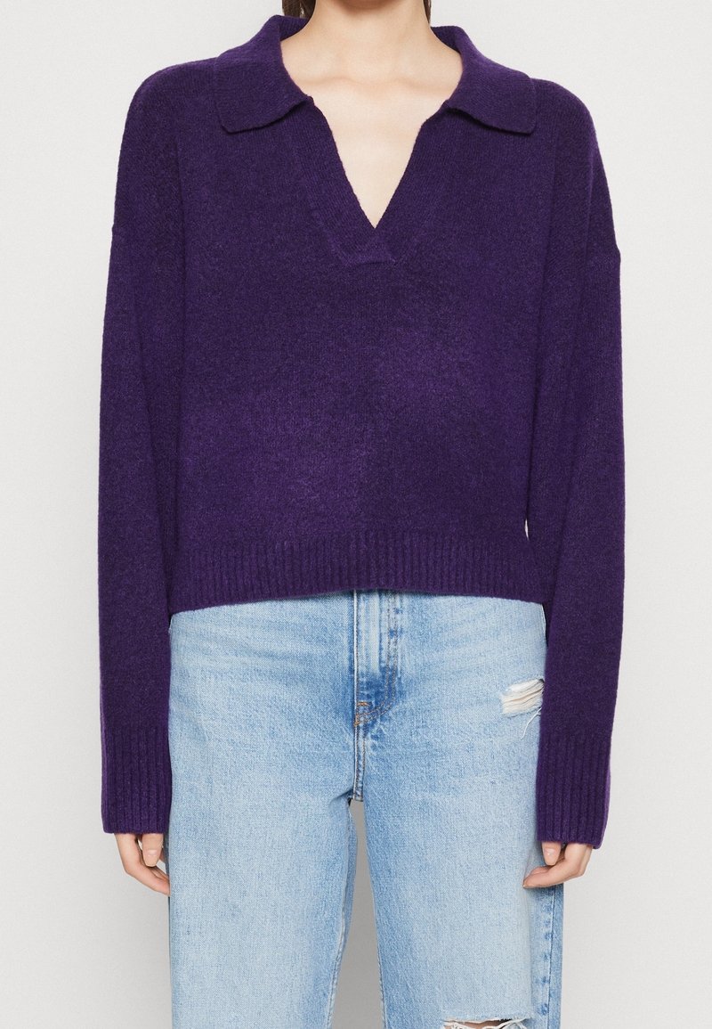 Lila stickad tröja med krage och V-ringning, med en avslappnad passform och ribbade ärmslut, ihop med ljusblå slitna jeans.